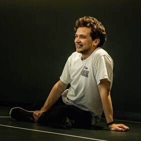 Joe Gilmour, white man aged 30, sits on floor of black studio theatre